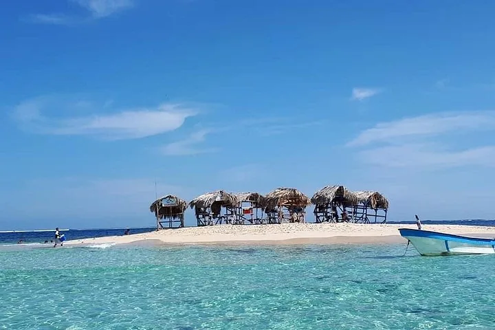 Small sandbar with several thatched huts and a boat in the foreground.
