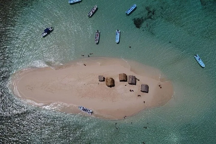Top-down aerial view of a sand island with huts and boats anchored around.